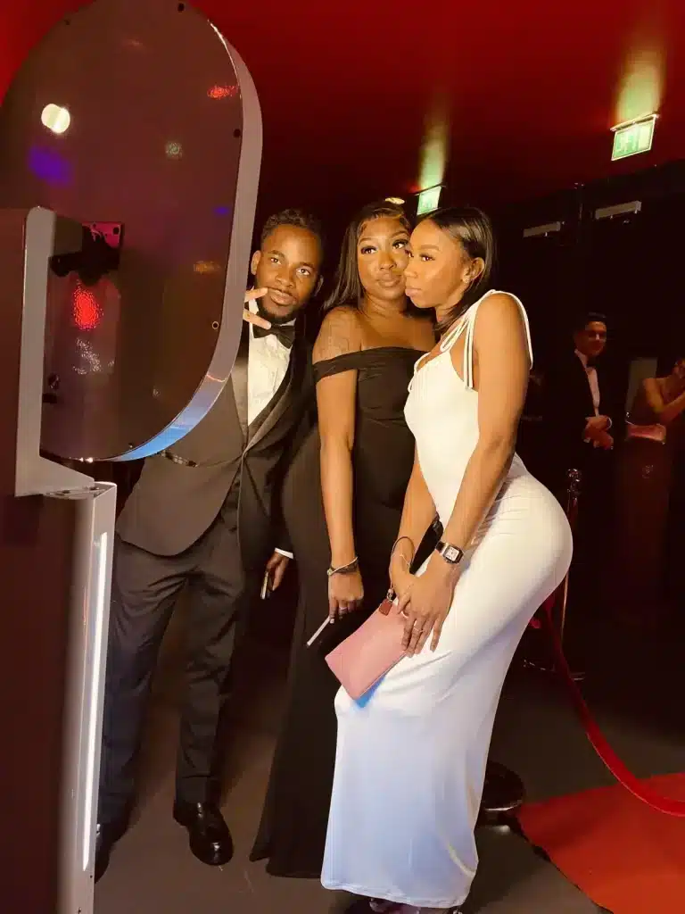 Three well-dressed people posing together in front of a photo booth at a formal event, standing on a red carpet with warm lighting.