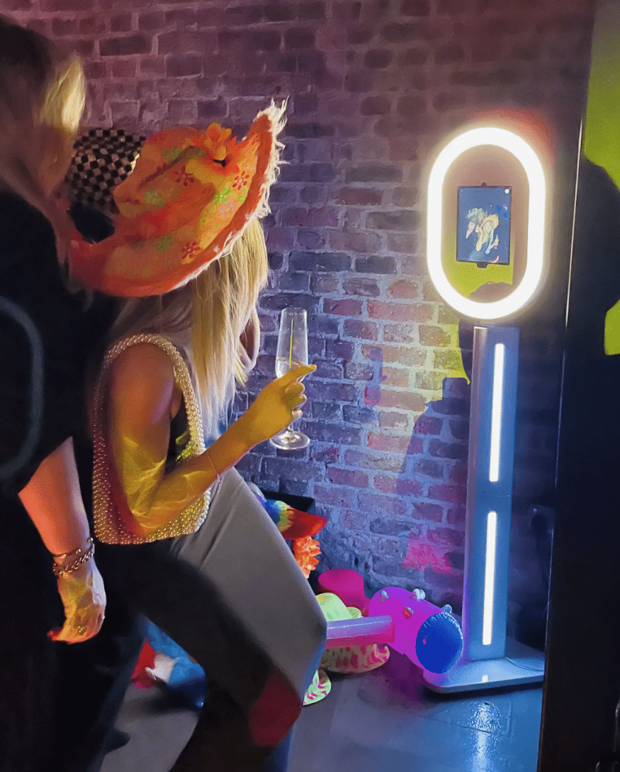 A group of smiling people gathered closely around a photo booth with a bright ring light, posing and laughing together at an indoor event.